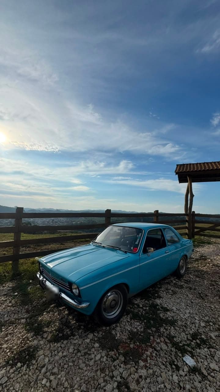 Cupê azul-claro antigo, de para-choque cromado e faróis redondos, parado num mirante no fim da tarde
