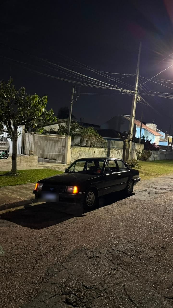 Cupê preto antigo visto de frente, parado na rua à noite, com os faróis de posição acesos