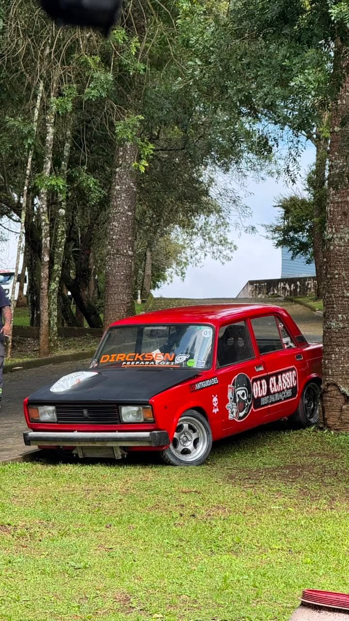 Lada Laika vermelho de capô preto, com faixa DIRCKSEN no para-brisa, parado na grama sob árvores