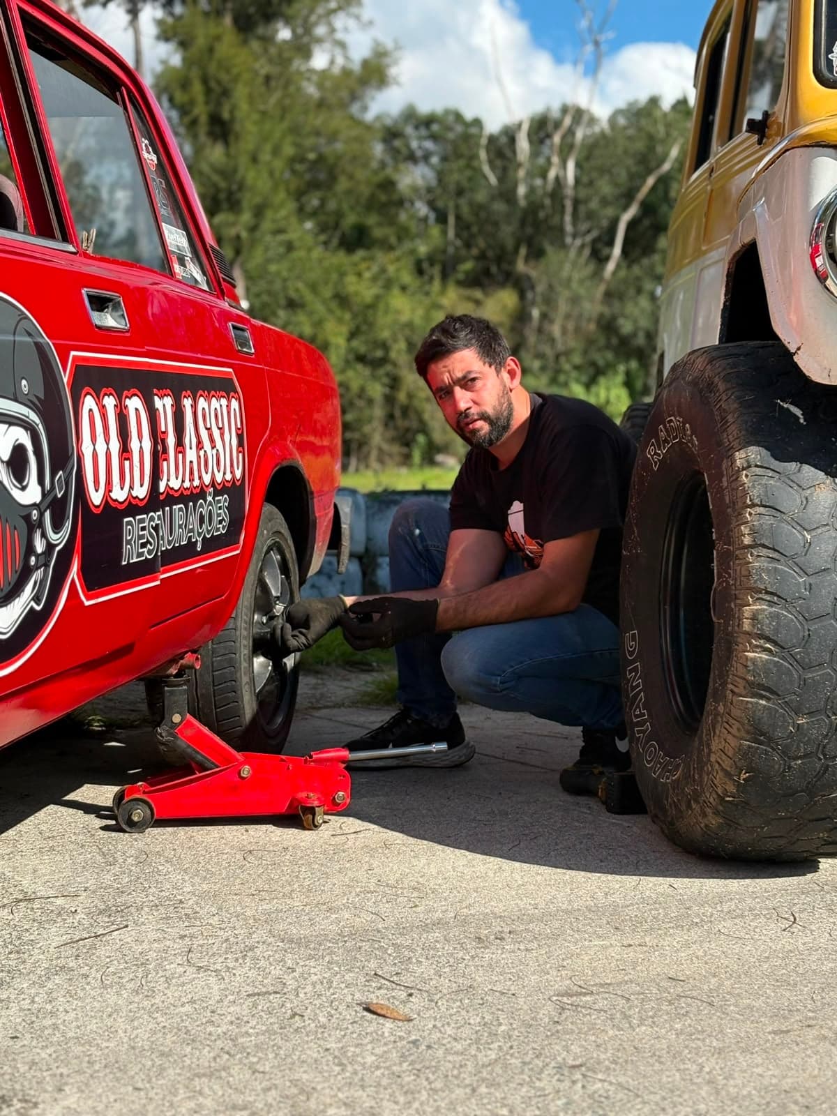 Victor Dircksen agachado trocando a roda dianteira de um Lada Laika vermelho erguido no macaco, com uma Rural amarela ao lado
